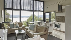 A modern living room with a large window, a sofa, a coffee table, and a kitchen area. - Tamar River Apartments (Grindelwald)
