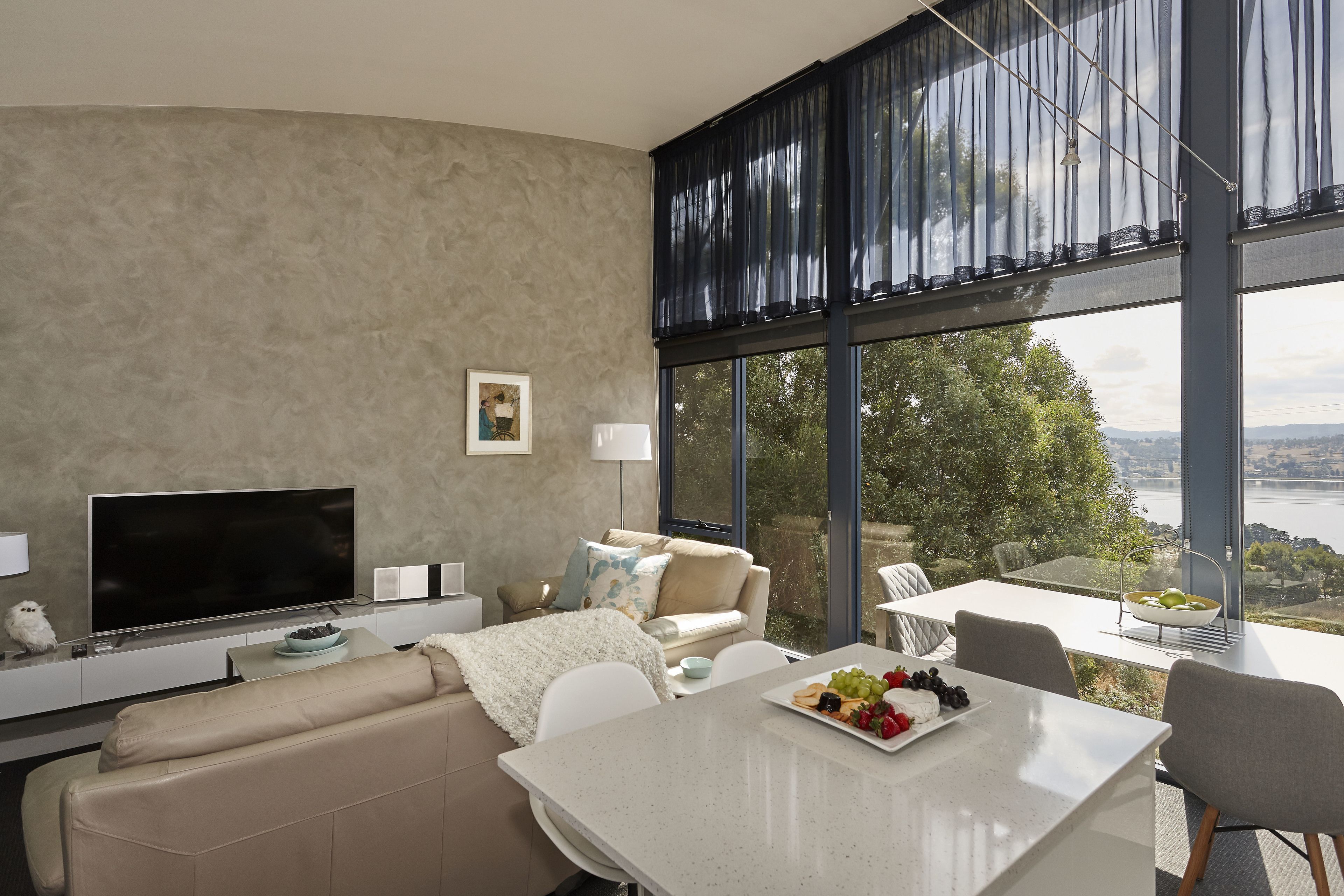 A modern living room with a large TV, a sofa, a dining table with fruit, and a view of a body of water.