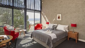 A modern bedroom with a large window, a bed with a grey bedspread, red chairs, and a small table. - Tamar River Apartments (Grindelwald)