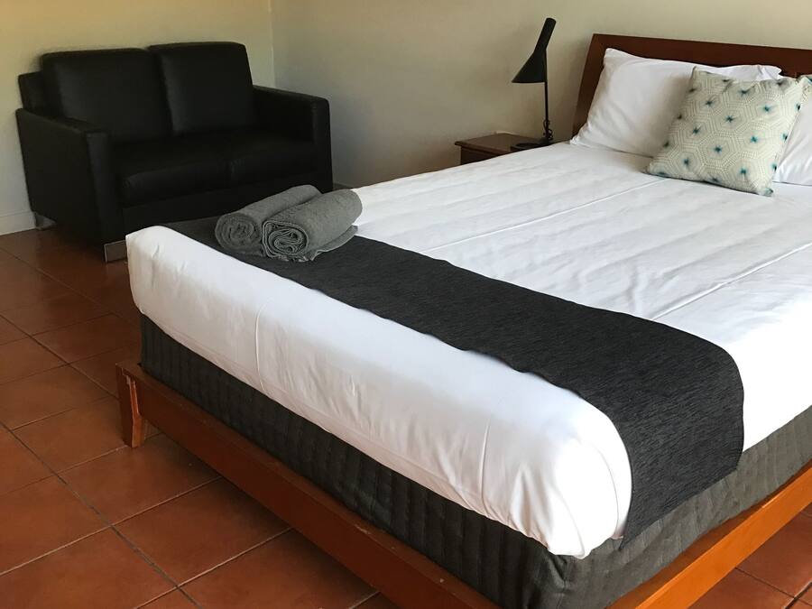 Un lit bien fait, avec une literie blanche et foncée, une chaise noire et une petite table de chevet avec une lampe.