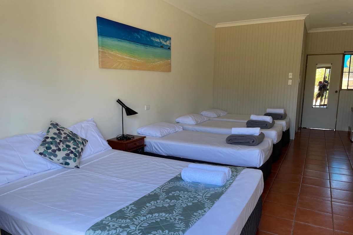 A hotel room with three beds, a painting of a beach, and a tiled floor.