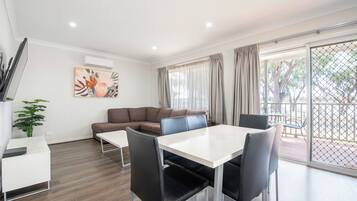 A modern living room with a sofa, a dining table with chairs, a potted plant, and a large window with curtains.