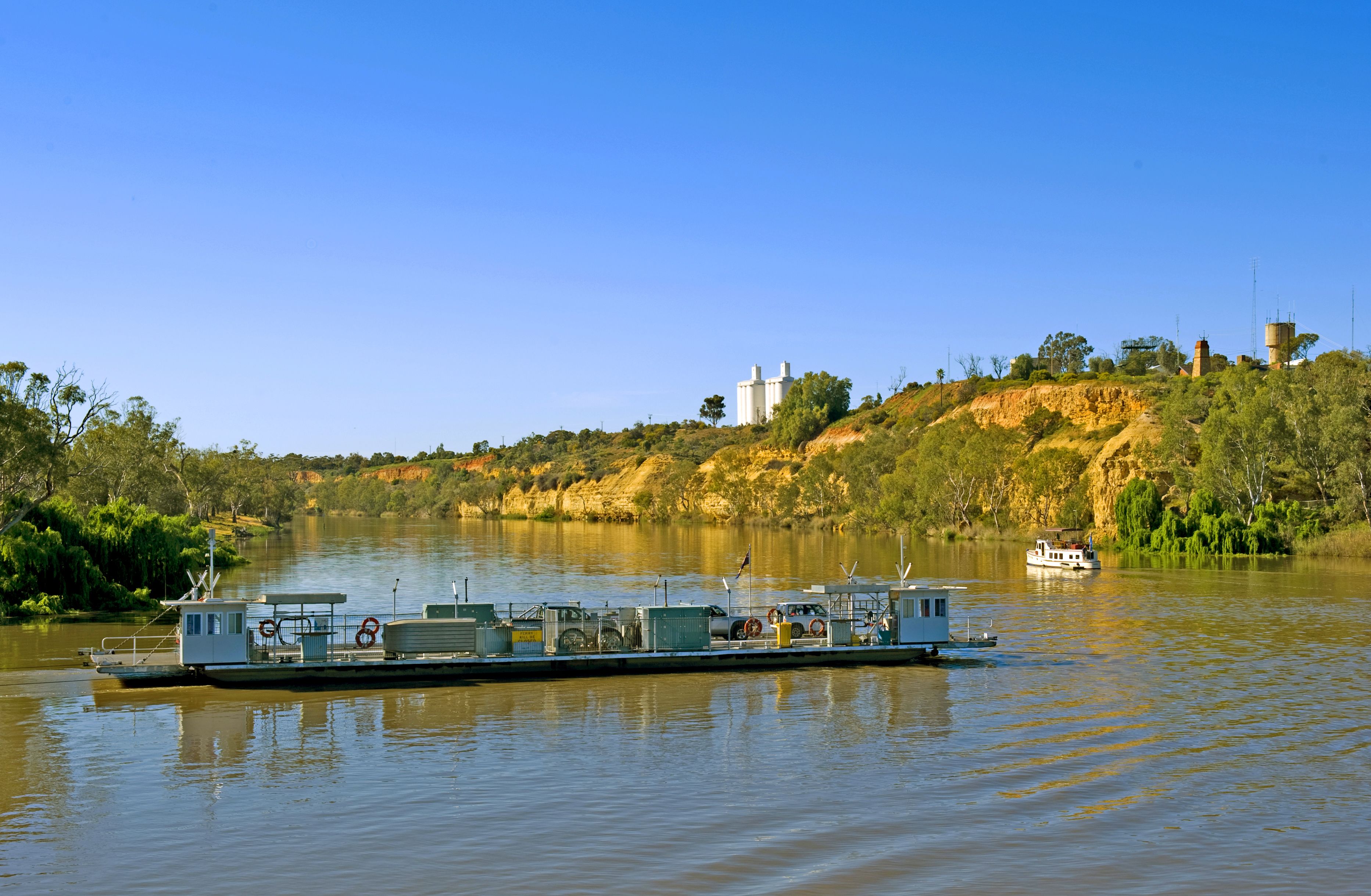 A riverboat with a flat deck and a small cabin, docked at a calm river with a hilly landscape in the background.