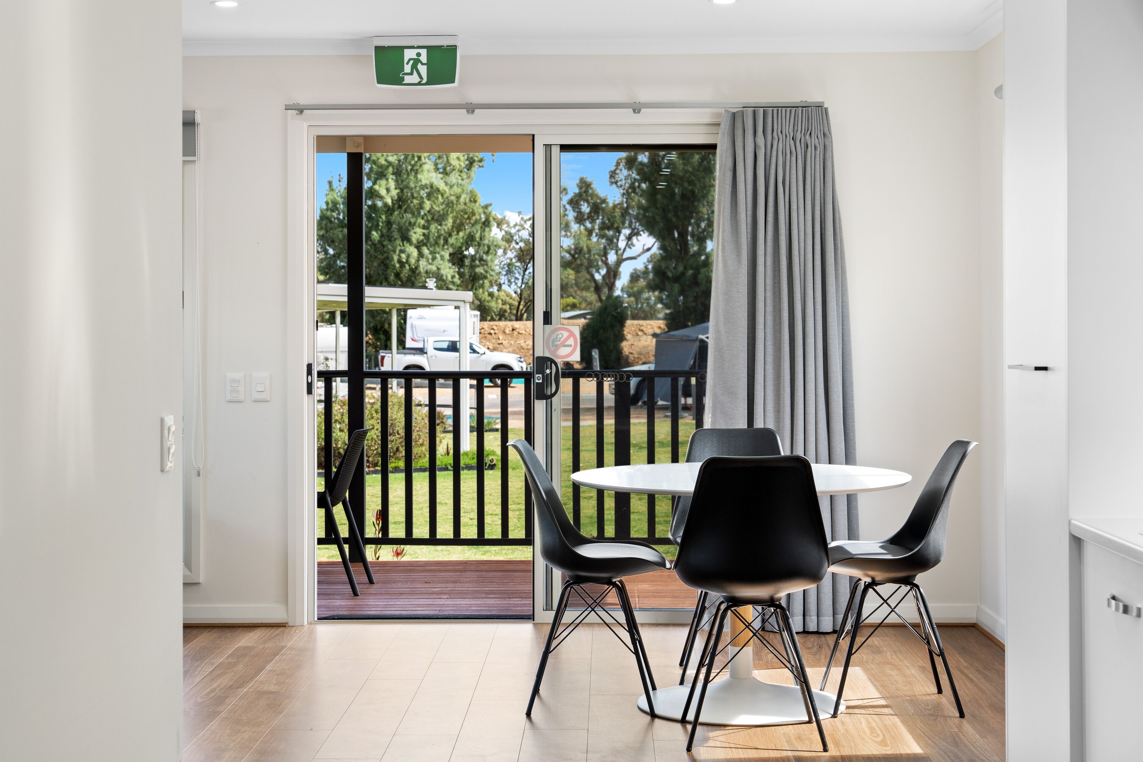 Une chambre moderne dotée d’une porte-fenêtre coulissante donnant sur un balcon, d’une table ronde avec quatre chaises et offrant une vue sur un camion blanc à l’extérieur.