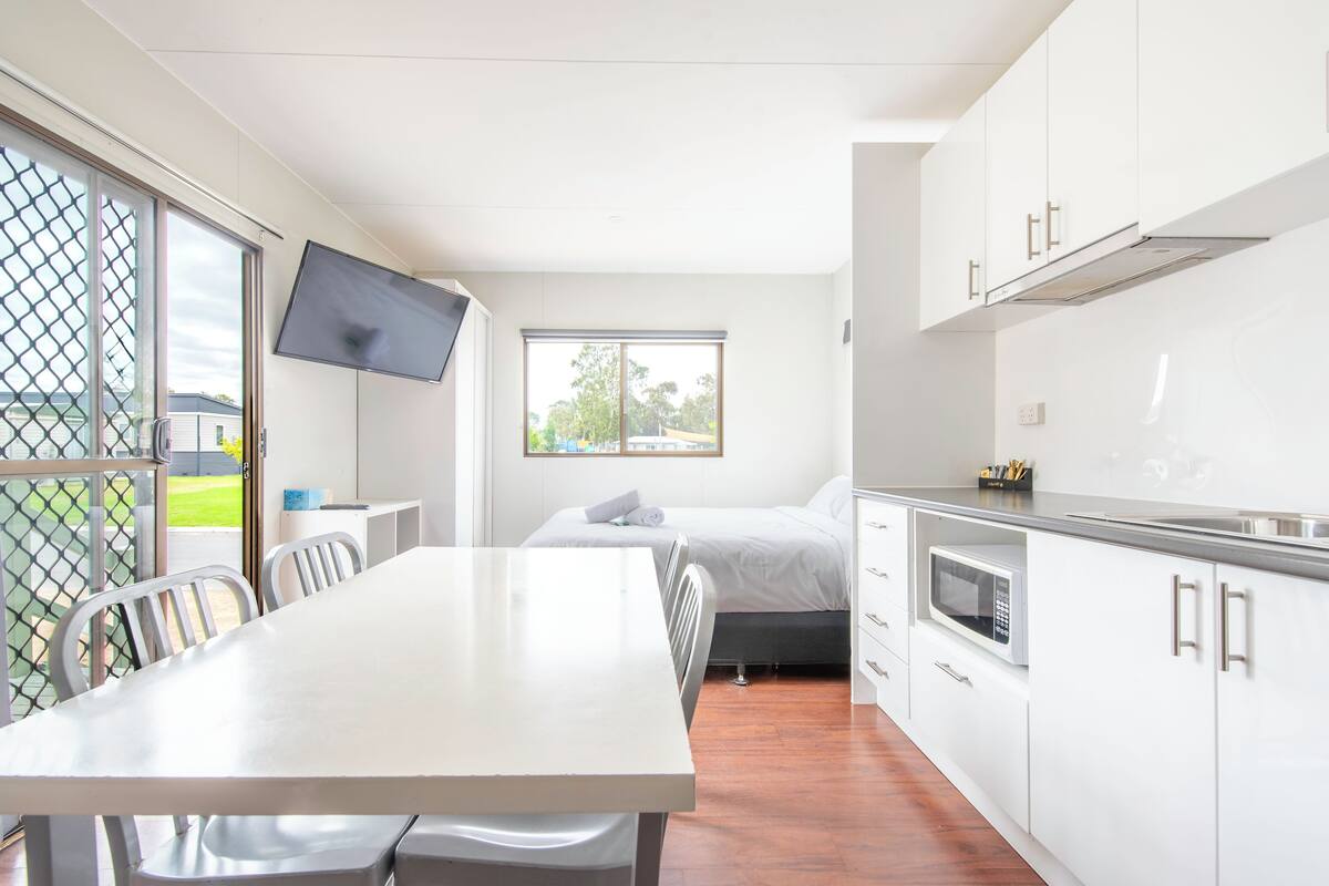 A modern, compact room with a dining table, a bed, a TV, and a kitchenette.