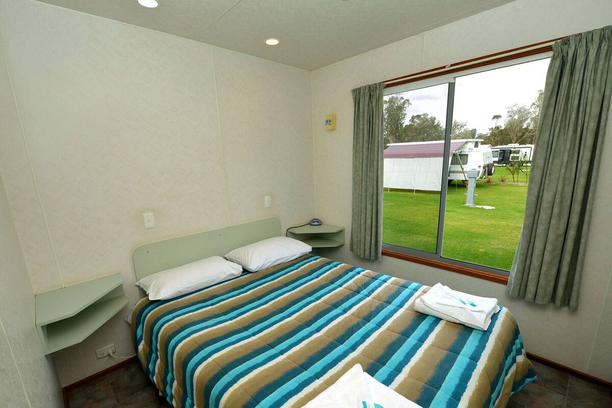 A single bed with a striped bedspread, a bedside table, a power outlet, and a window with curtains.