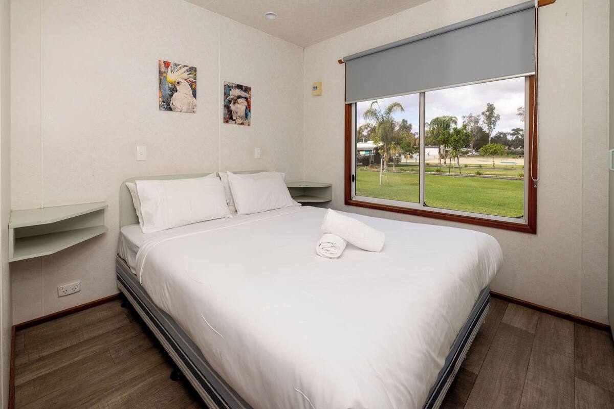 A hotel room with a bed, a window with a view of a grassy area, and two wall-mounted artworks.