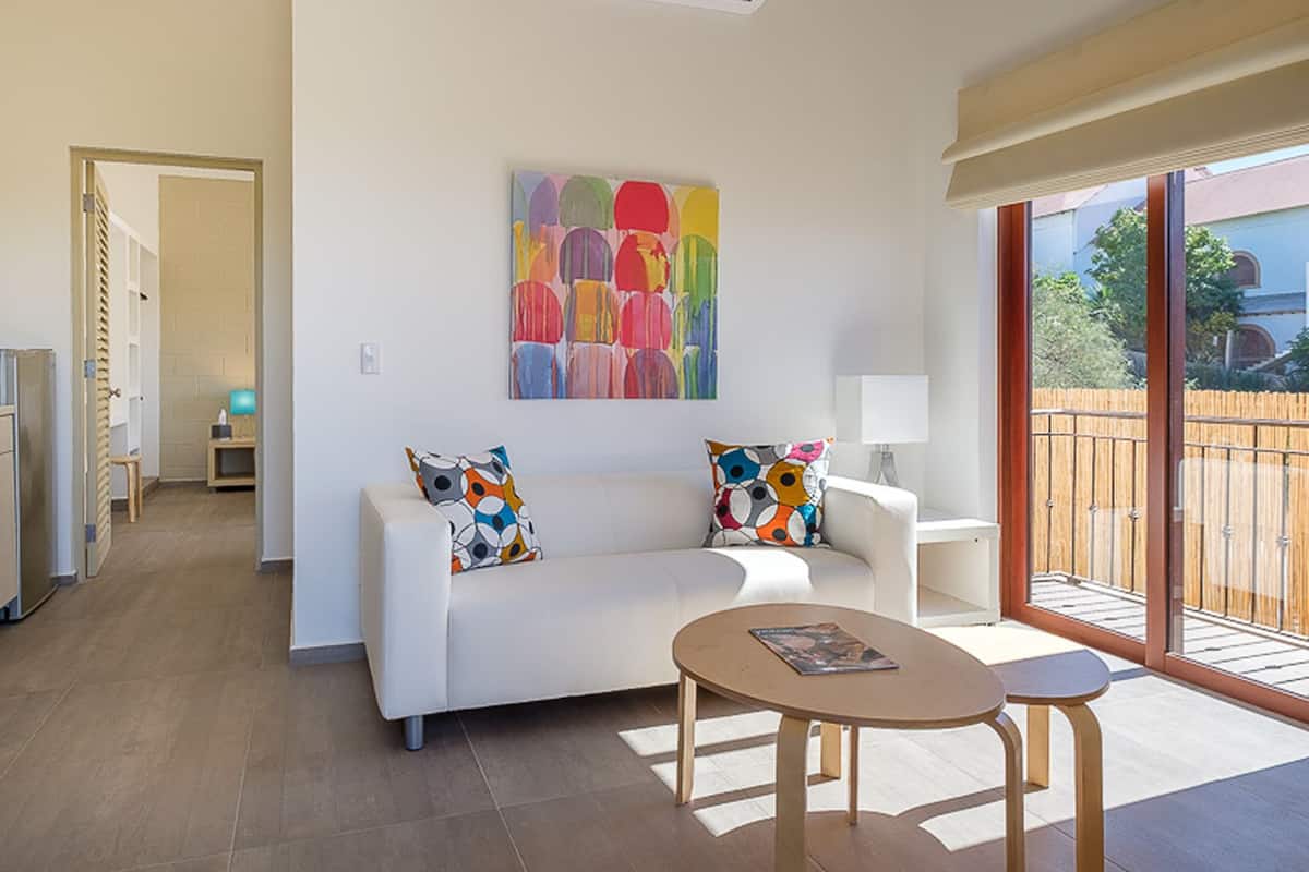 Una sala de estar moderna con un sofá blanco, cojines de colores, una pequeña mesa de madera y un ventanal con vistas al exterior.