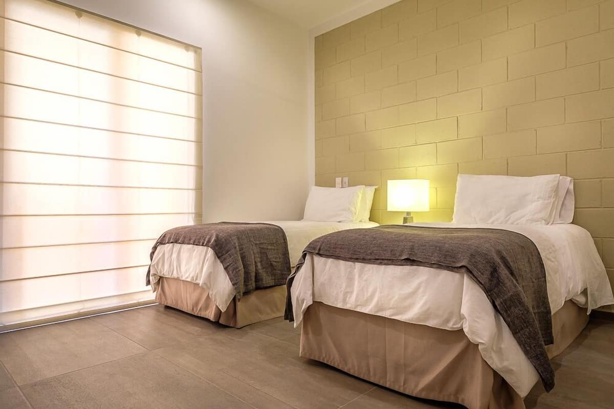 Una habitación de hotel con dos camas, una pared de ladrillo, una ventana con persianas y una lámpara de noche.