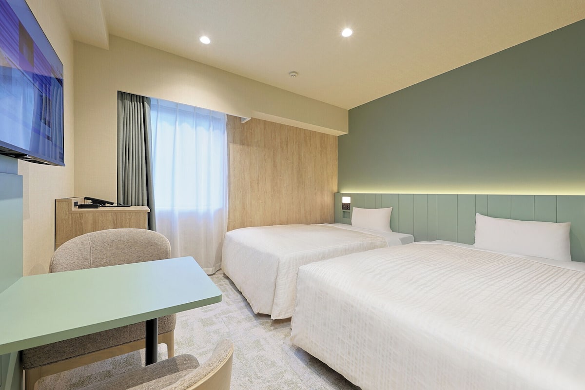 A hotel room with two beds, a desk, a chair, a TV, and a window with curtains.