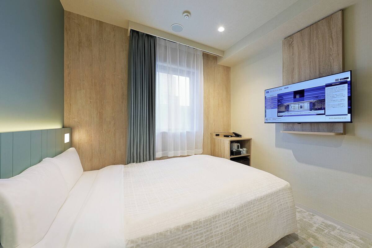 A hotel room with a bed, a TV mounted on the wall, and a small shelf with a phone and a cup.