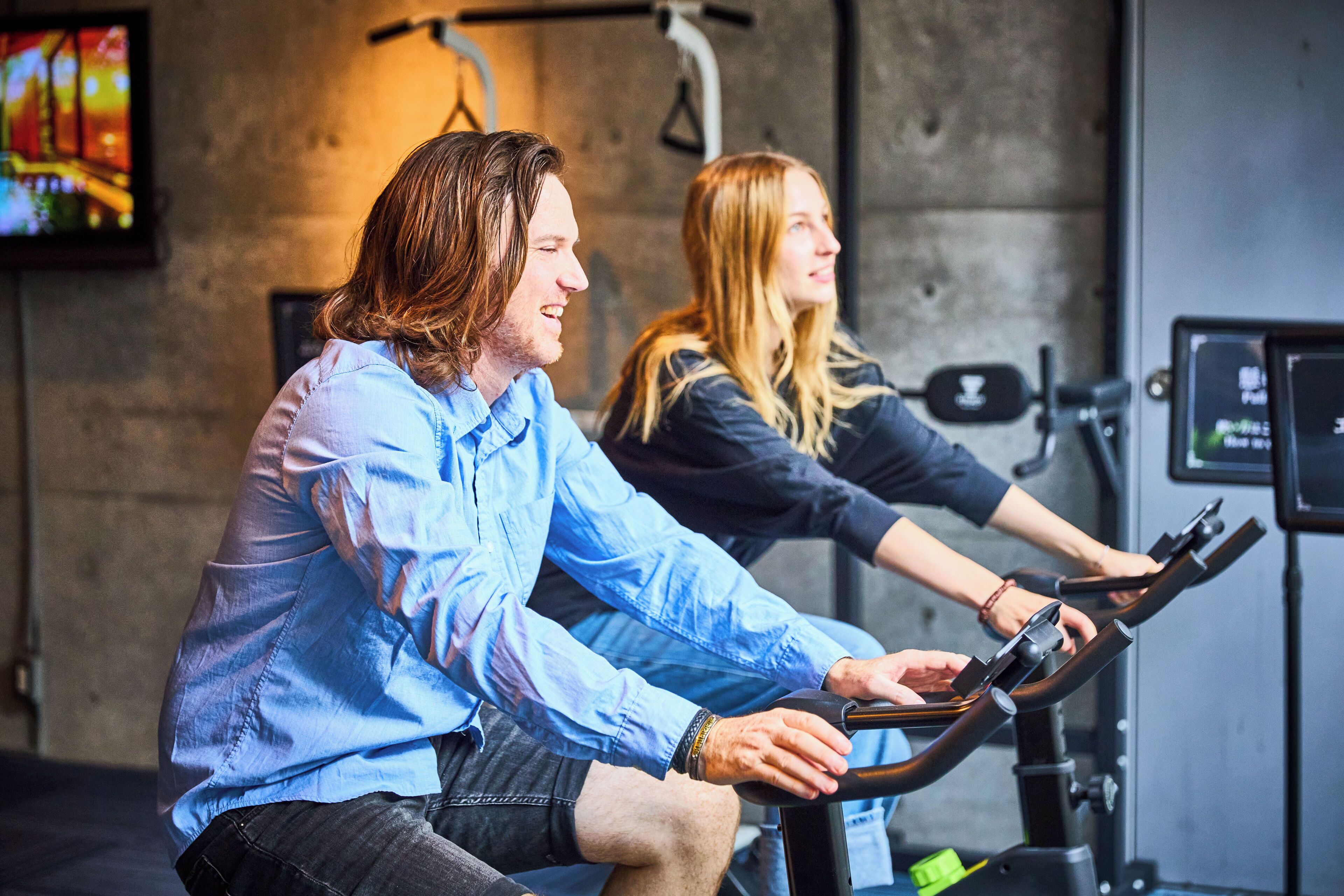 Two people using stationary bikes in a gym with a concrete wall and a TV screen in the background.