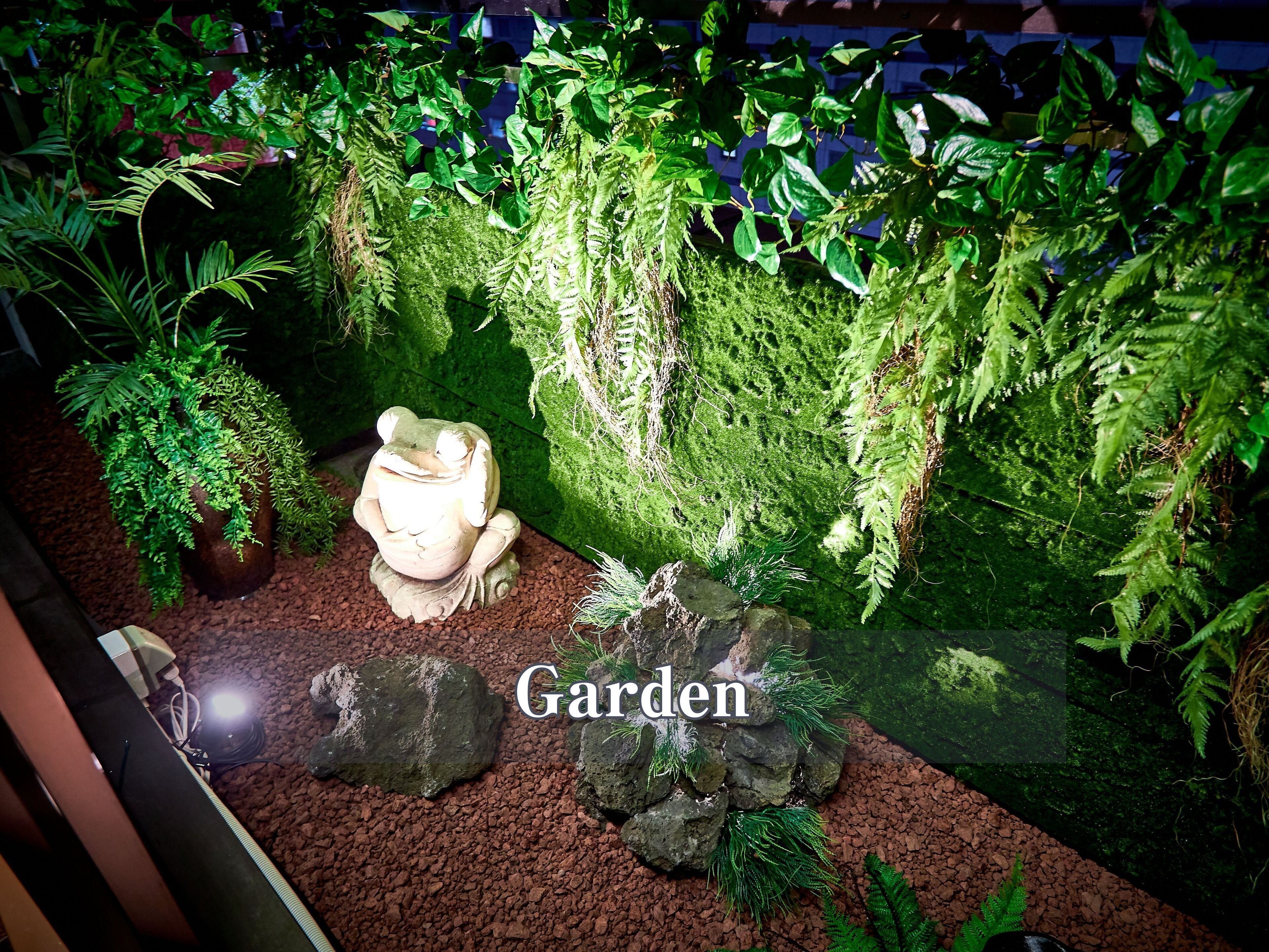 Un jardín con una estatua de rana, rocas y diversas plantas.