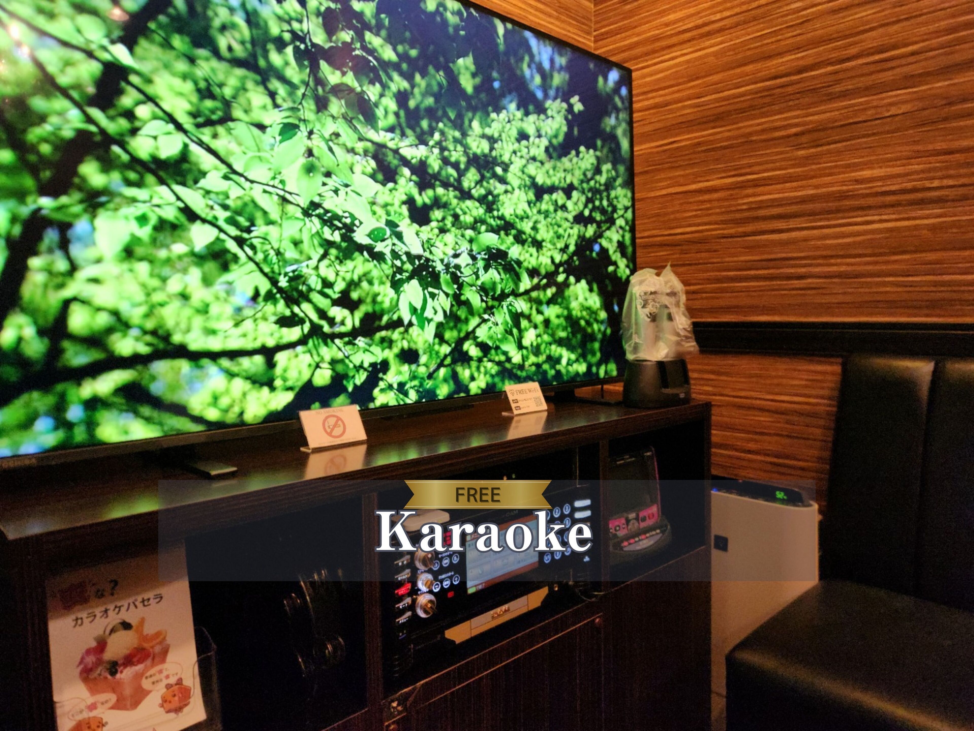 A karaoke room with a large TV displaying a forest scene, a coffee machine, and a sign indicating free karaoke.