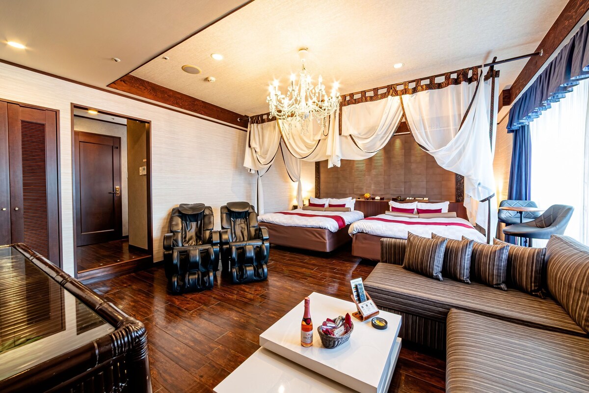 Une chambre d’hôtel avec un grand lit, un canapé, des fauteuils massants, une table basse avec une bouteille et un panier, et un lustre.
