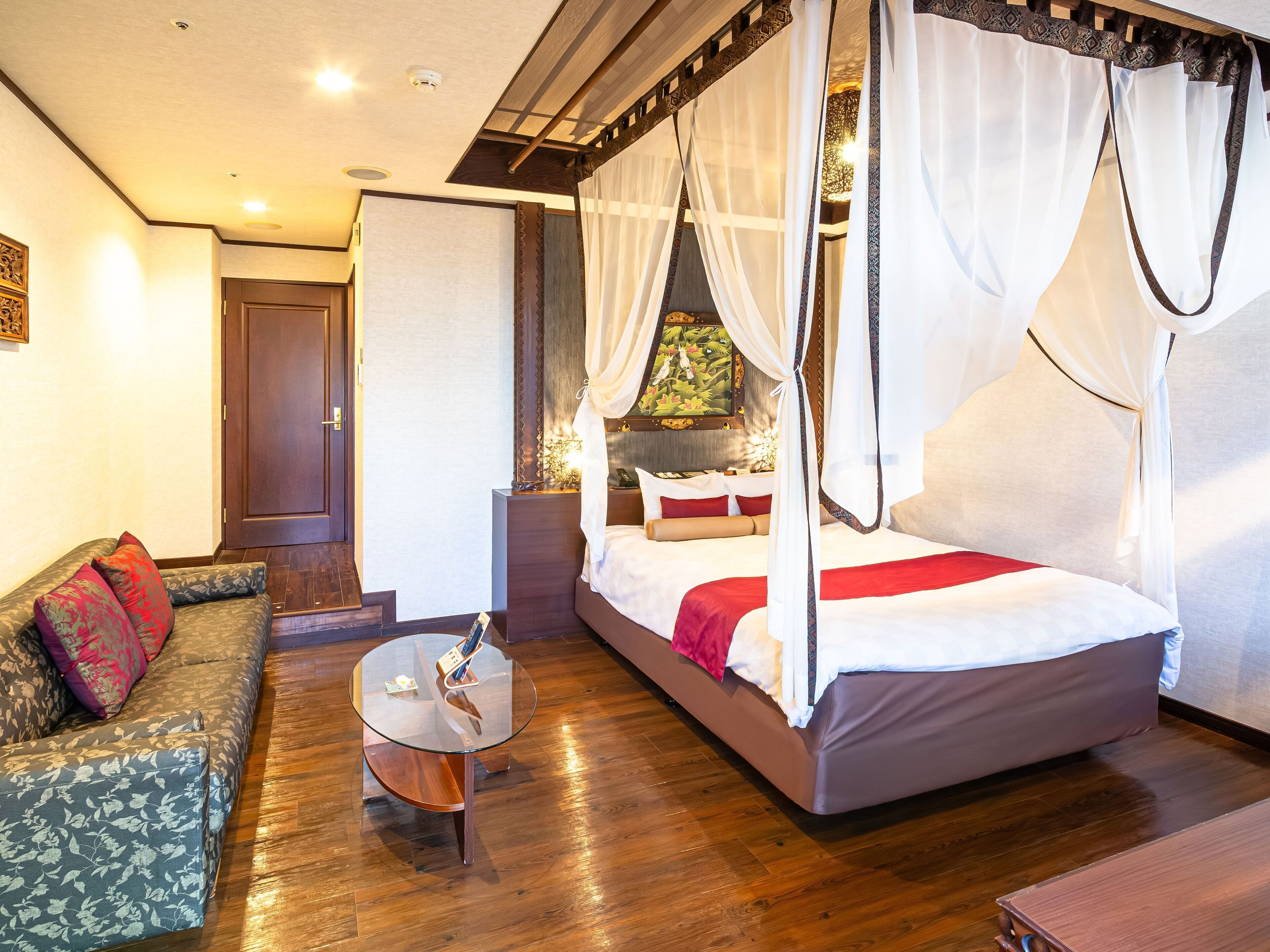 A bedroom with a four-poster bed, a sofa, a glass coffee table, and a wooden floor.