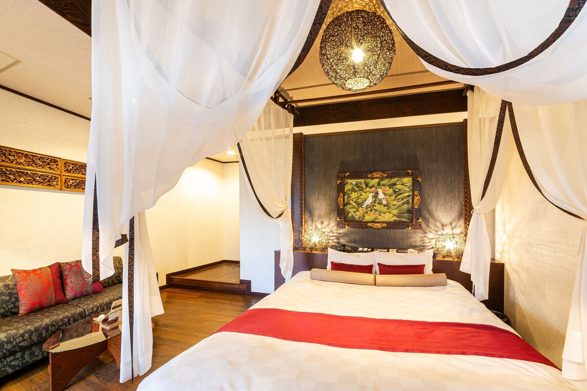 Une chambre à coucher avec un lit à baldaquin, un canapé avec des coussins rouges, une table basse en bois et un panneau décoratif mural.