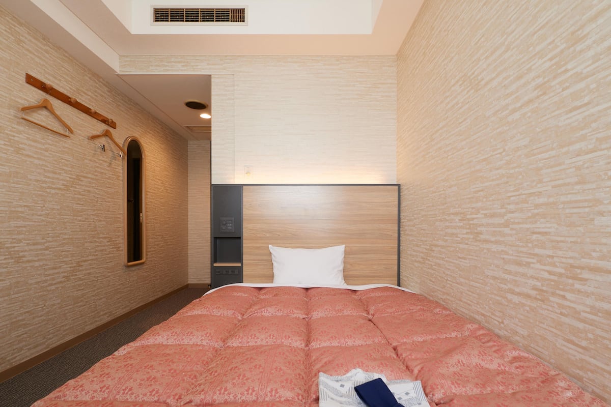 A hotel room with a bed, a wooden headboard, a wall-mounted heater, and a small window.
