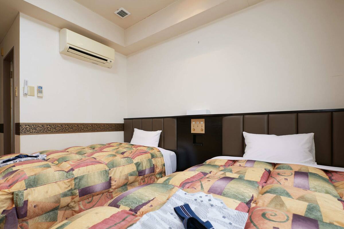 A hotel room with two beds, an air conditioning unit, a wall-mounted phone, and a control panel.