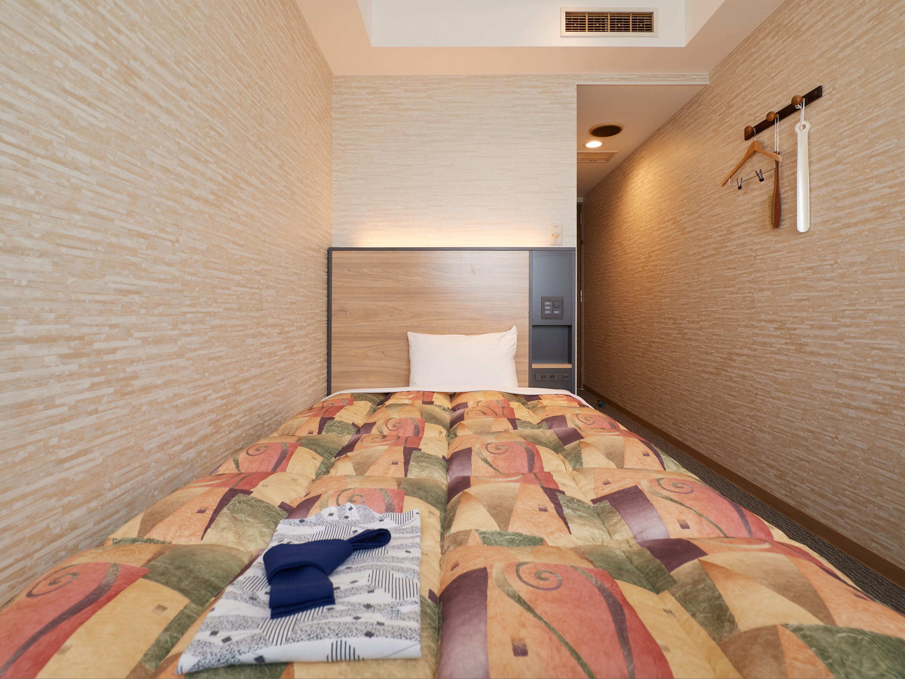 A narrow hotel room with a patterned bedspread, a neatly folded towel, and a hanging coat rack.