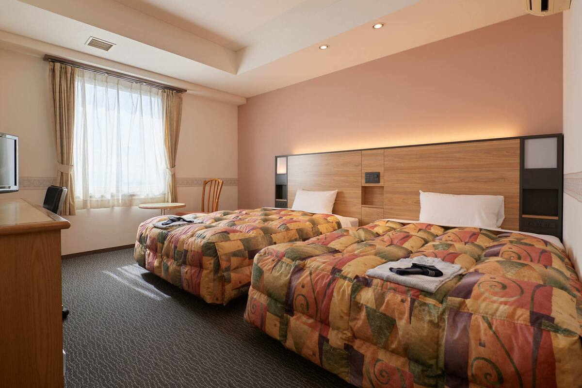 A hotel room with two beds, a desk, a chair, a TV, and a window with curtains.