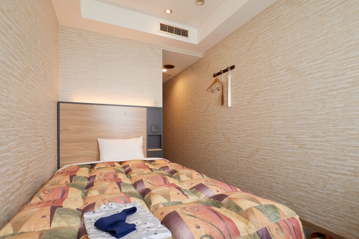 A hotel room with a bed, a headboard, a bedside table, a hanging coat rack, and a pair of slippers.