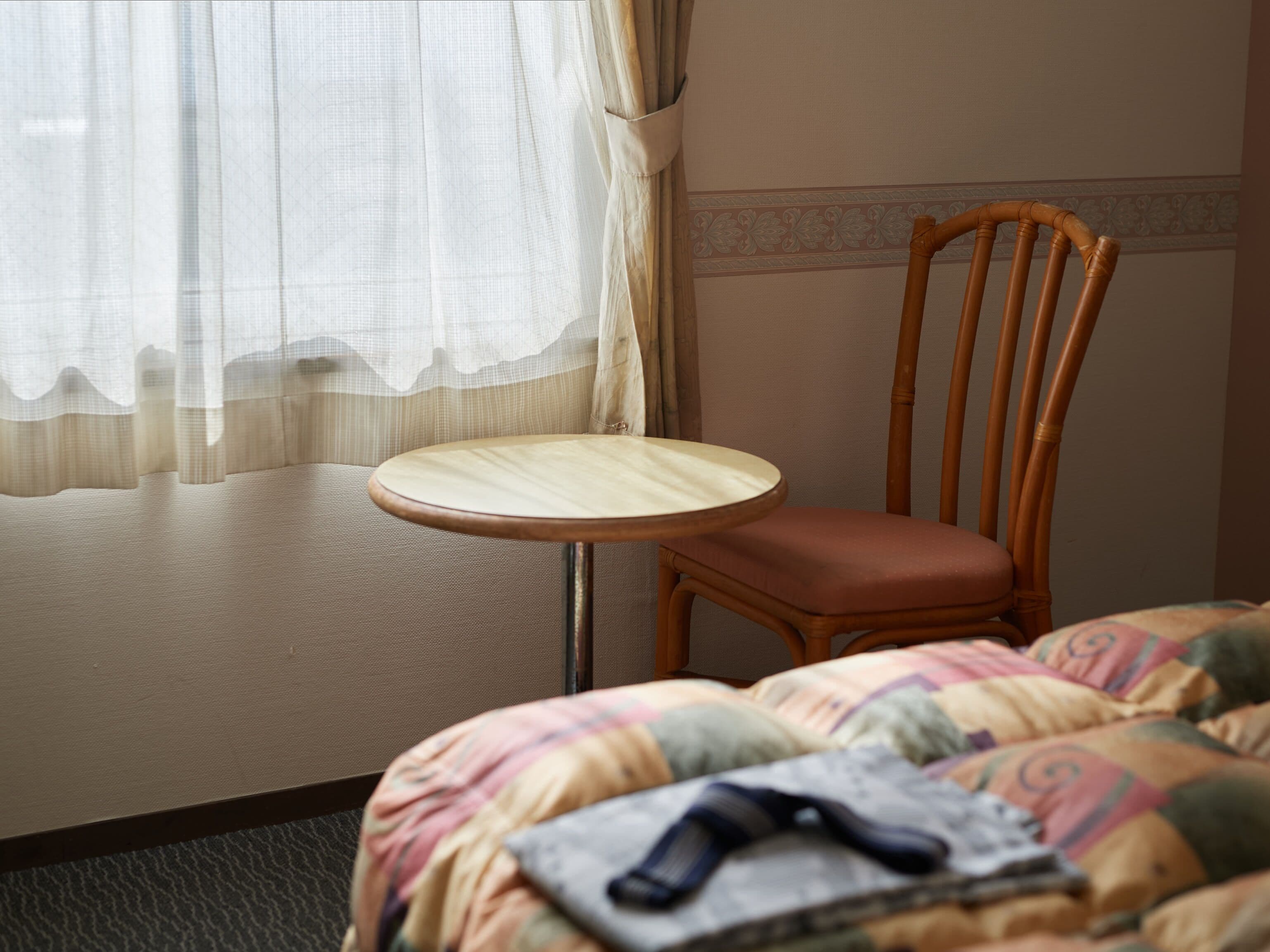 A hotel room with a bed, a small round table, a wooden chair, and a window with sheer curtains.