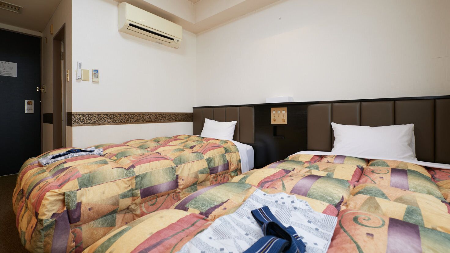 A hotel room with two beds, an air conditioning unit, a door, and a wall-mounted phone.
