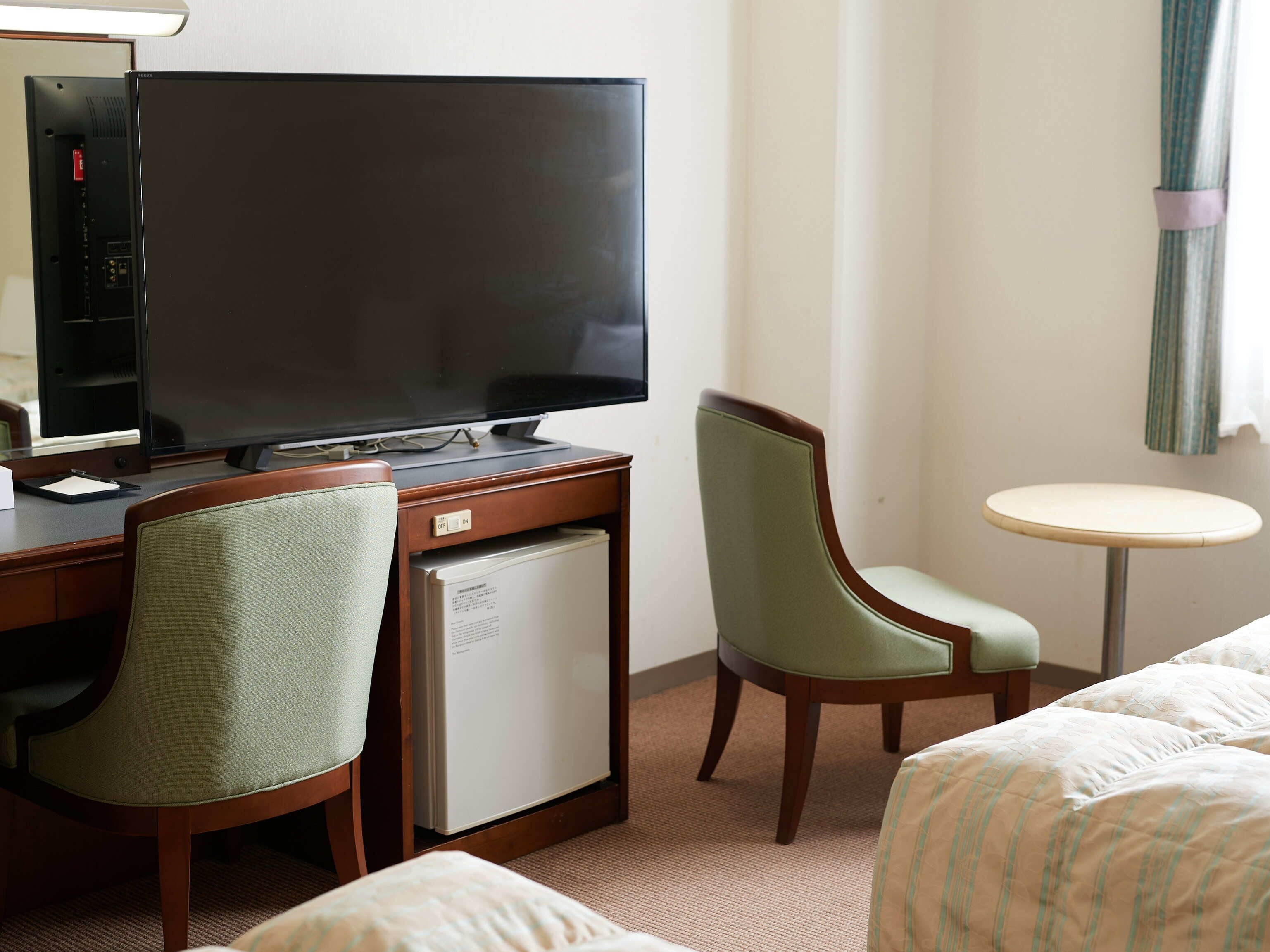 A hotel room with a flat-screen TV, a desk with a computer, a small fridge, two chairs, a bed, and a round table.