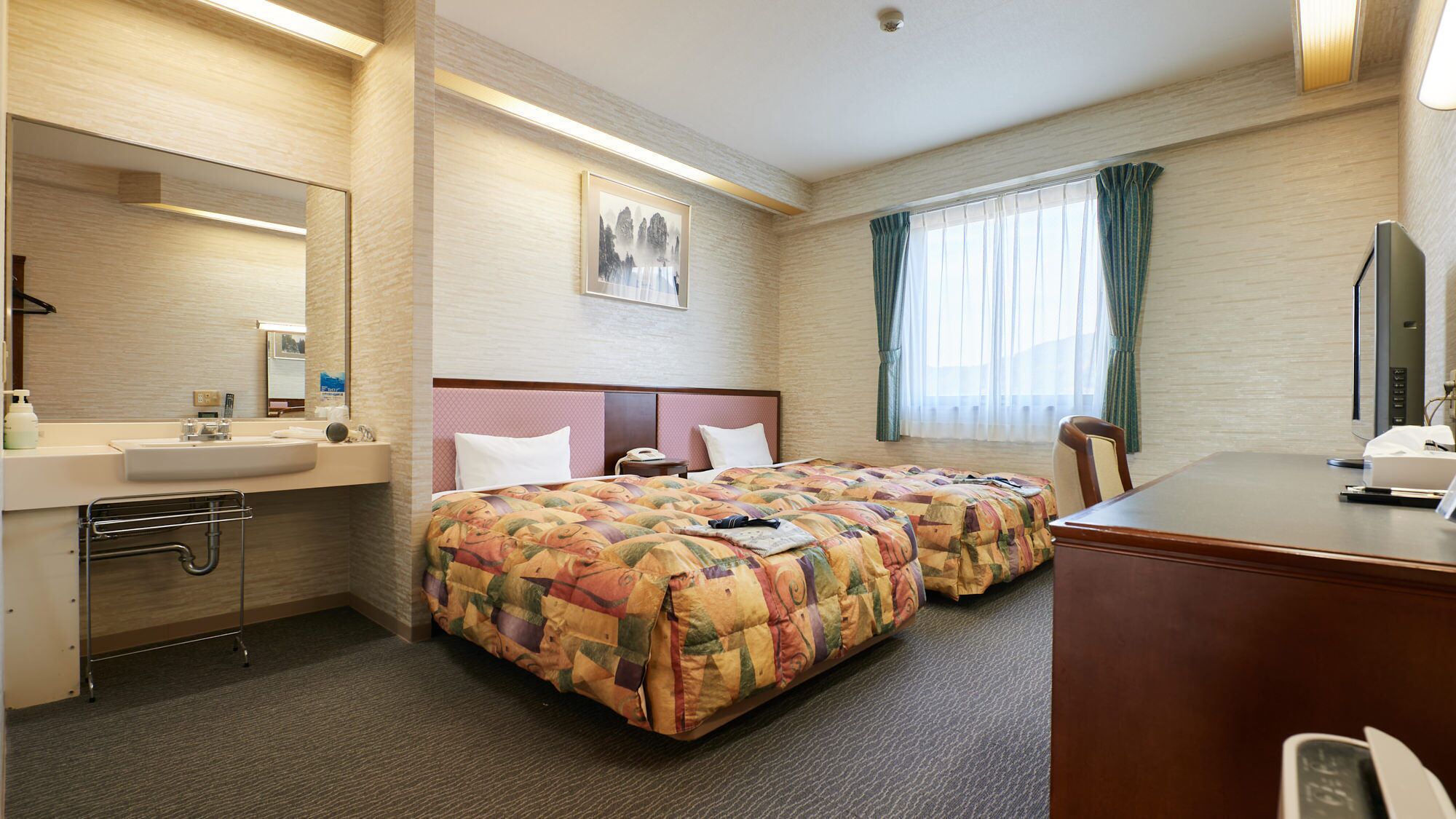 A hotel room with a bed, a desk, a chair, a desk lamp, a mirror, a sink, and a window with curtains.