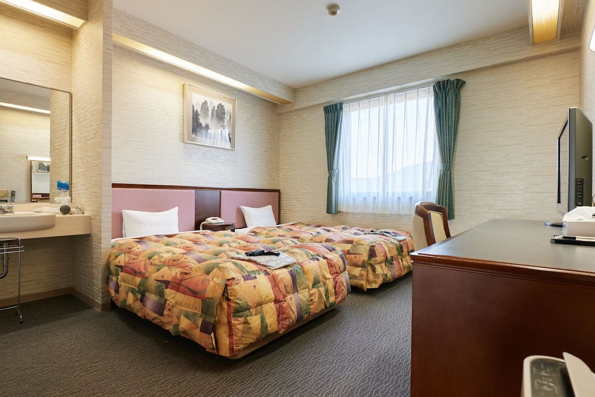 A hotel room with a bed, a desk, a chair, a desk lamp, a mirror, a sink, and a window with curtains.