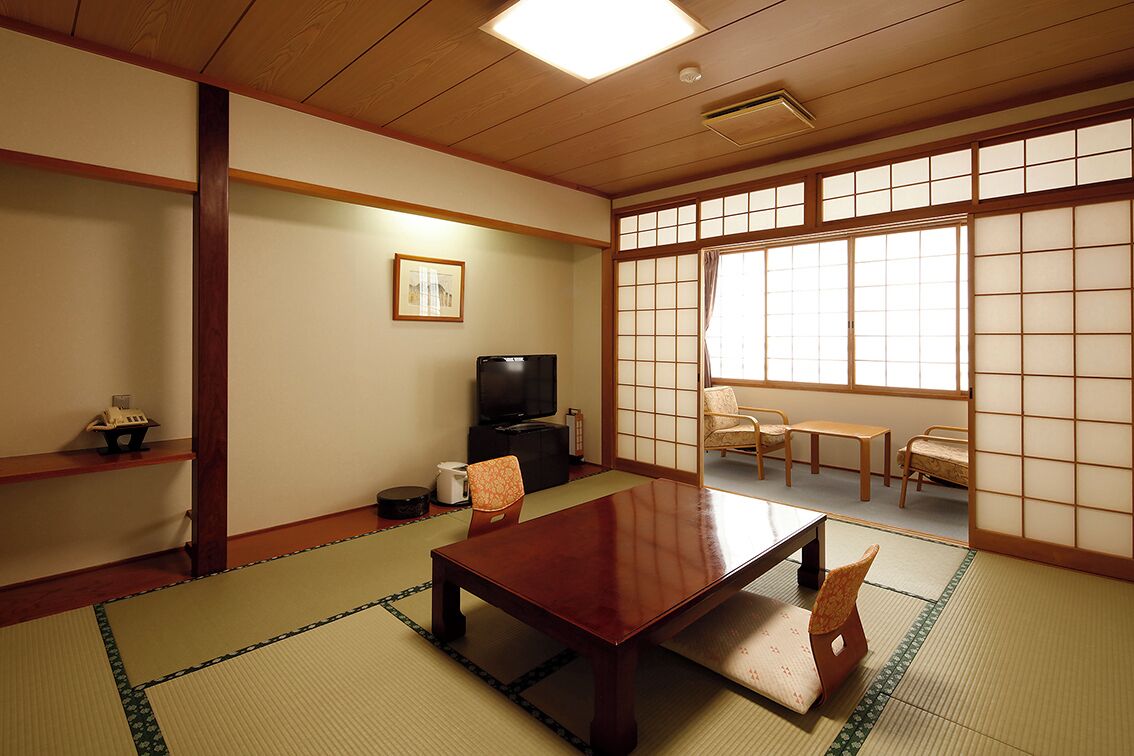 Une pièce de style japonais traditionnel, avec un sol recouvert de tatamis, une table basse, des chaises, un téléviseur et des portes coulissantes.