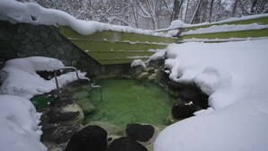 Un manantial de aguas termales al aire libre cubierto de nieve, rodeado de rocas y un muro de piedra.