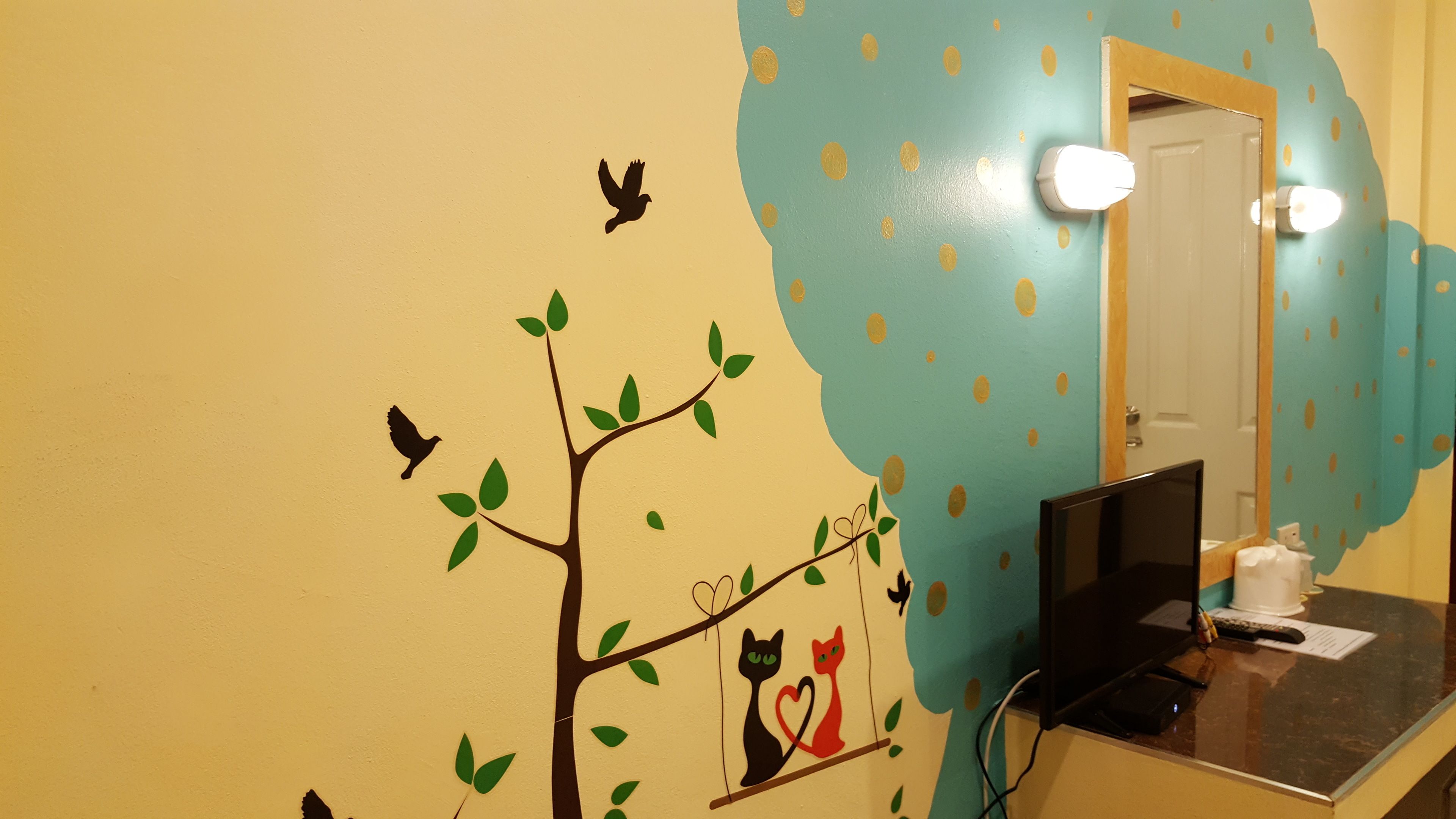 Une salle de bain ornée d’une fresque murale représentant un arbre, des oiseaux et des chats. On y trouve un miroir avec des lumières, un lavabo et des toilettes.