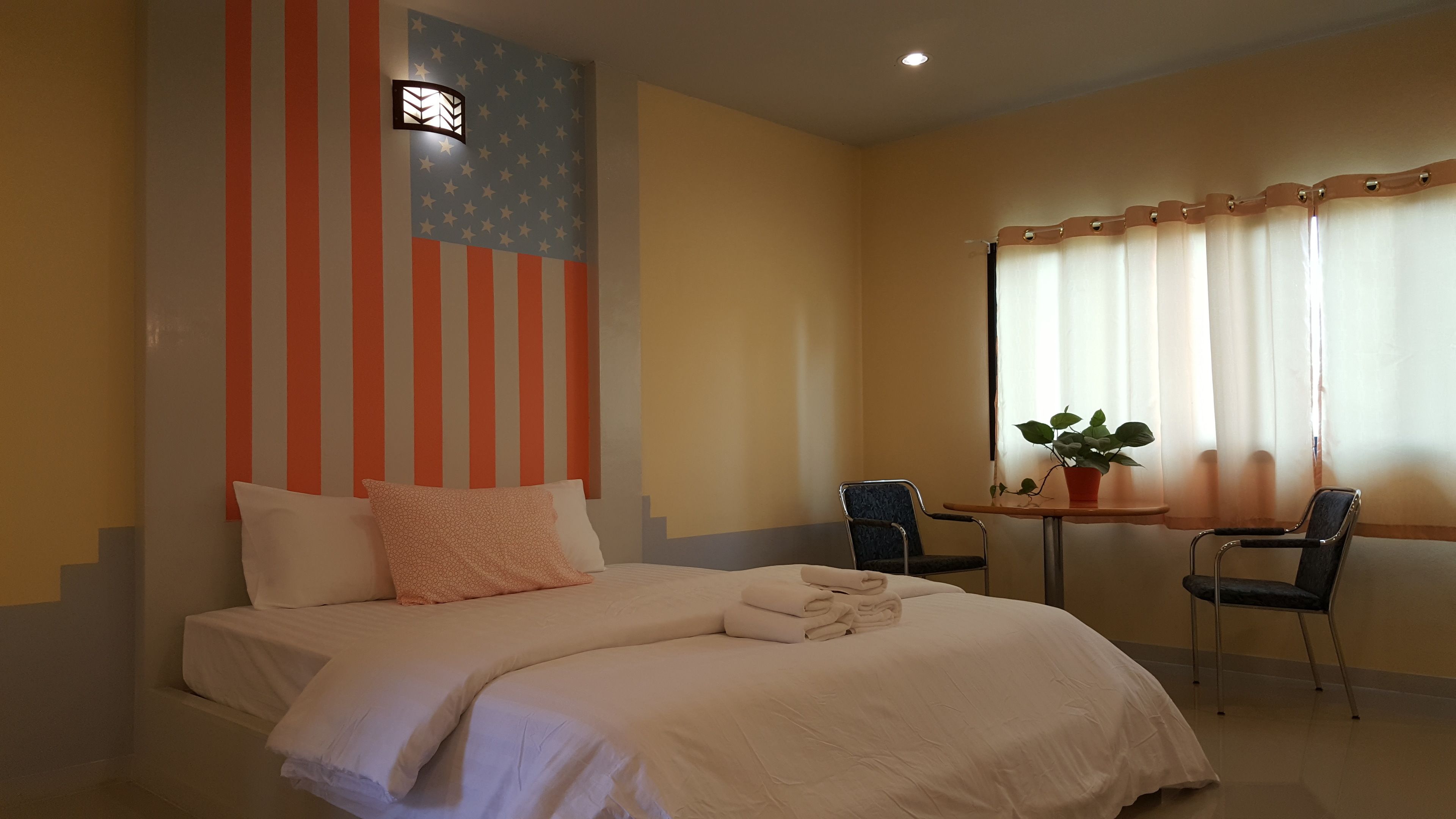 A hotel room with a bed, two chairs, a small table, a potted plant, and a window with curtains.