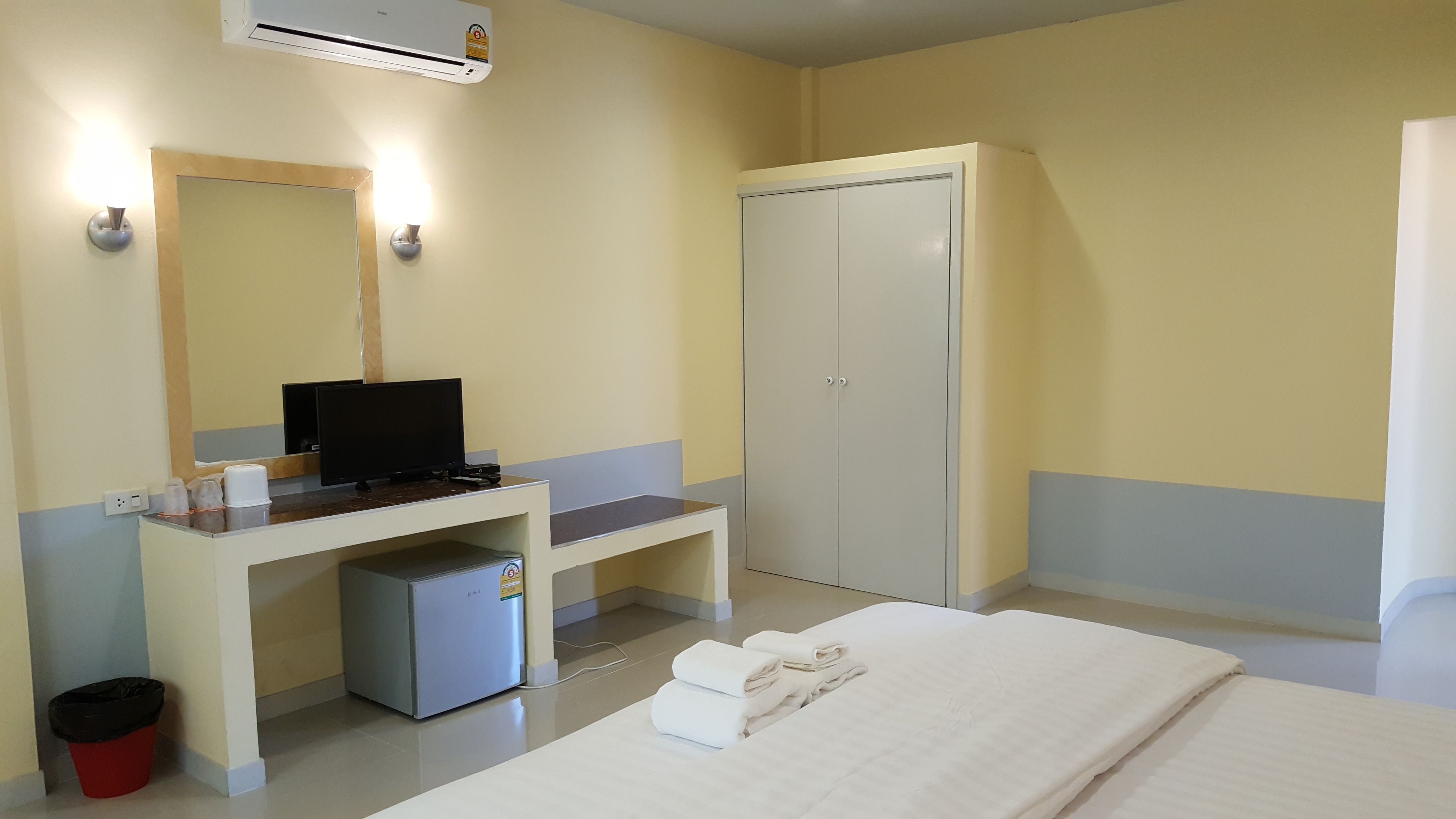 A hotel room with a bed, a desk with a computer, a small refrigerator, a wardrobe, and a trash bin.