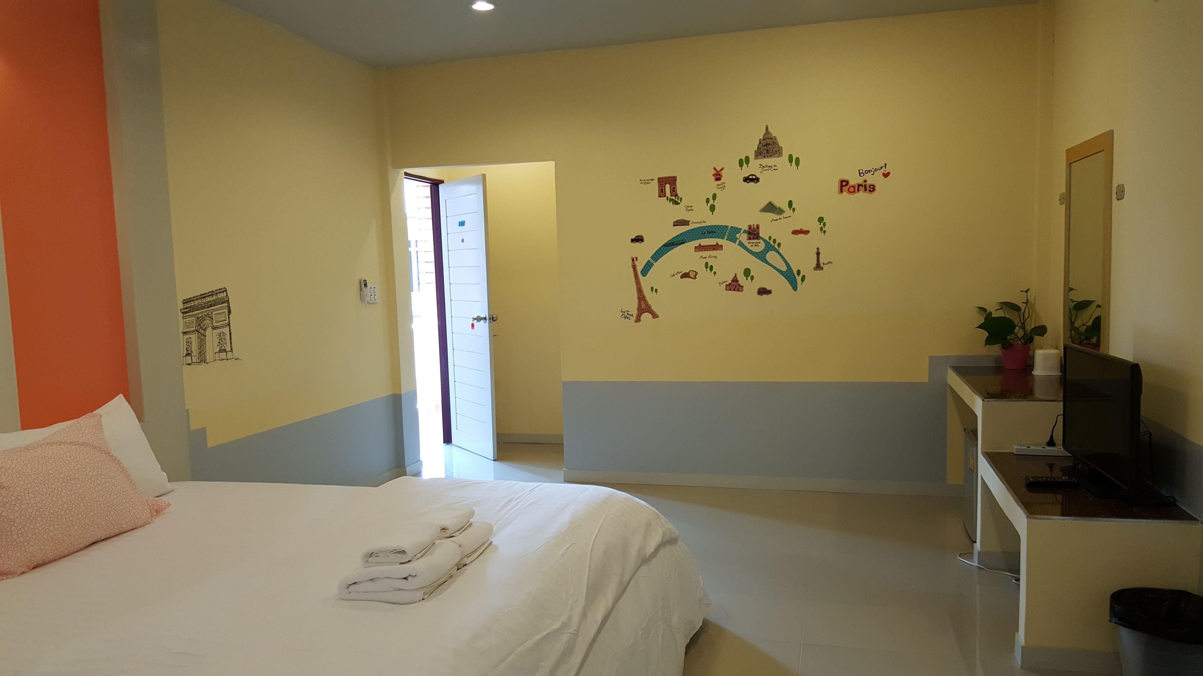 A hotel room with a bed, a desk with a computer, a wall with Paris-themed decorations, and a door leading to another room.