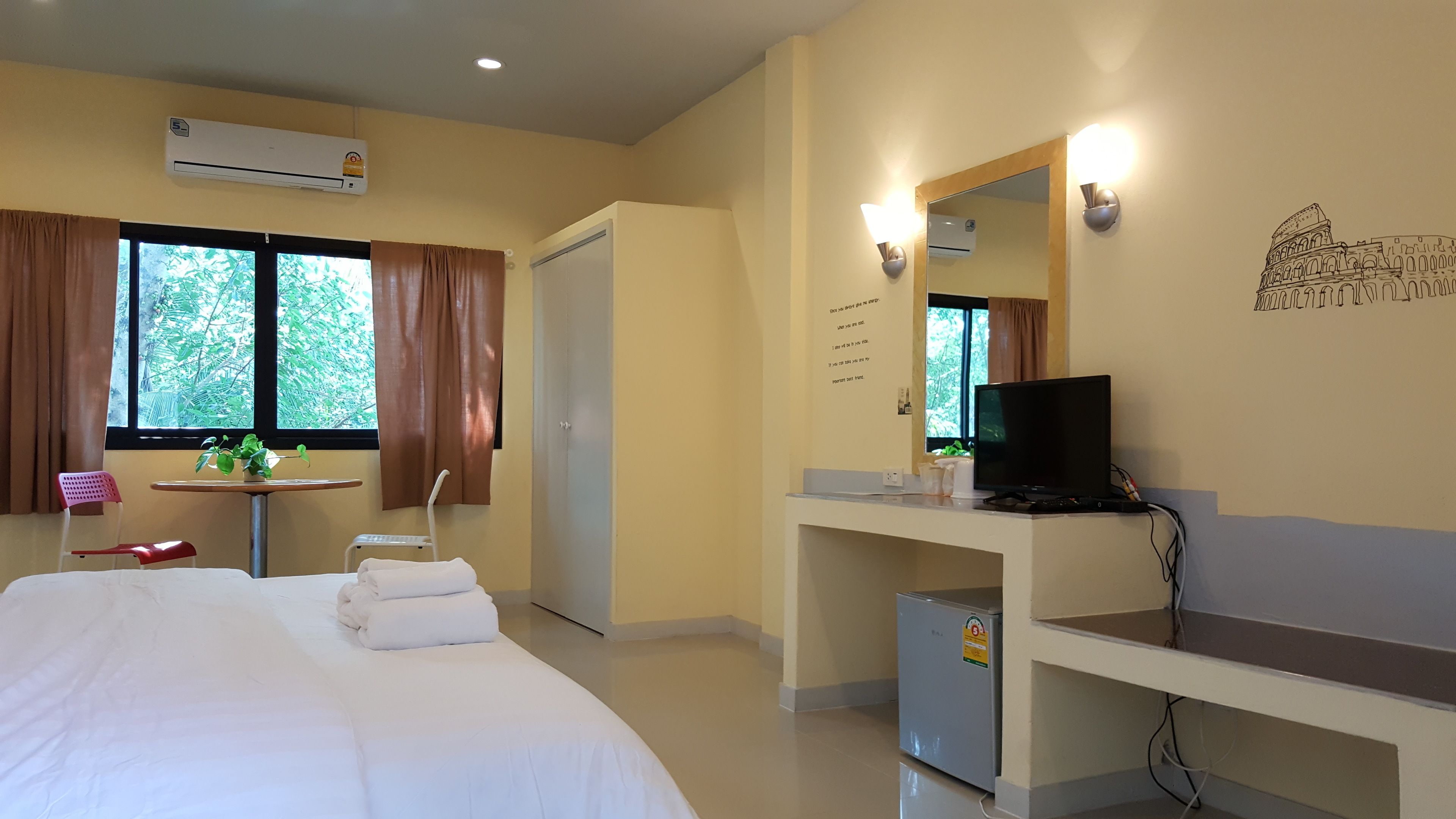 A hotel room with a bed, a desk with a computer, a small fridge, a window with curtains, and an air conditioning unit.