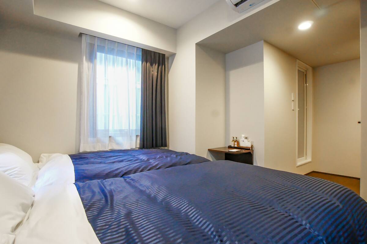 A hotel room with two beds, a small bedside table, an air conditioning unit, and a window with curtains.