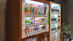 A vending machine with various drinks and snacks, a small display of hotel room images, and a plant with a red ribbon. - Hotel Livemax Tokyo Bakurocho (Tokyo)