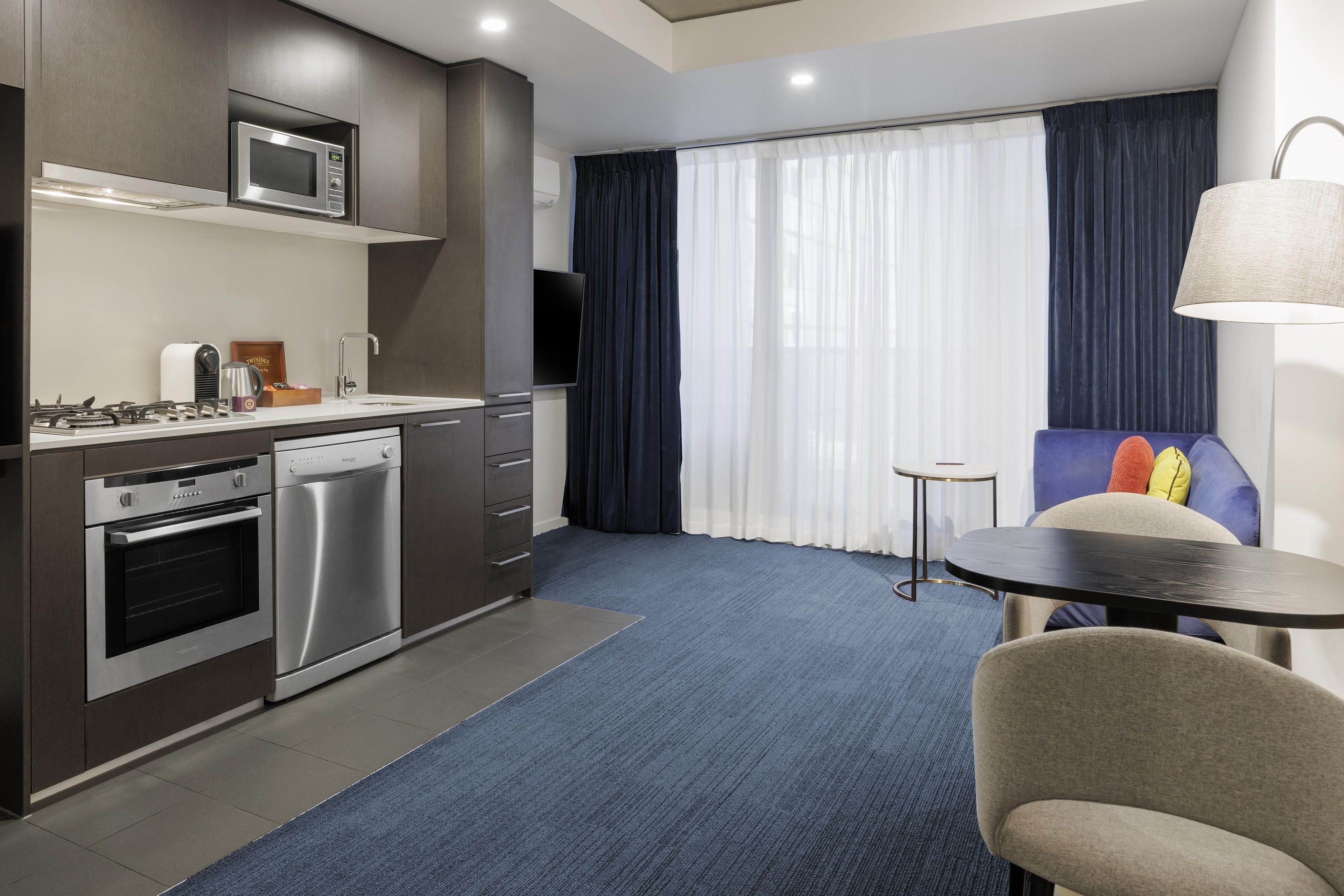 A compact hotel room with a kitchenette, a dining area, and a large window with curtains.