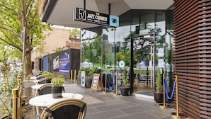 Uma área de estar ao ar livre com cadeiras e mesas de vime, uma placa indicando The Jazz Corner Hotel, e uma entrada de vidro com vista para a rua.