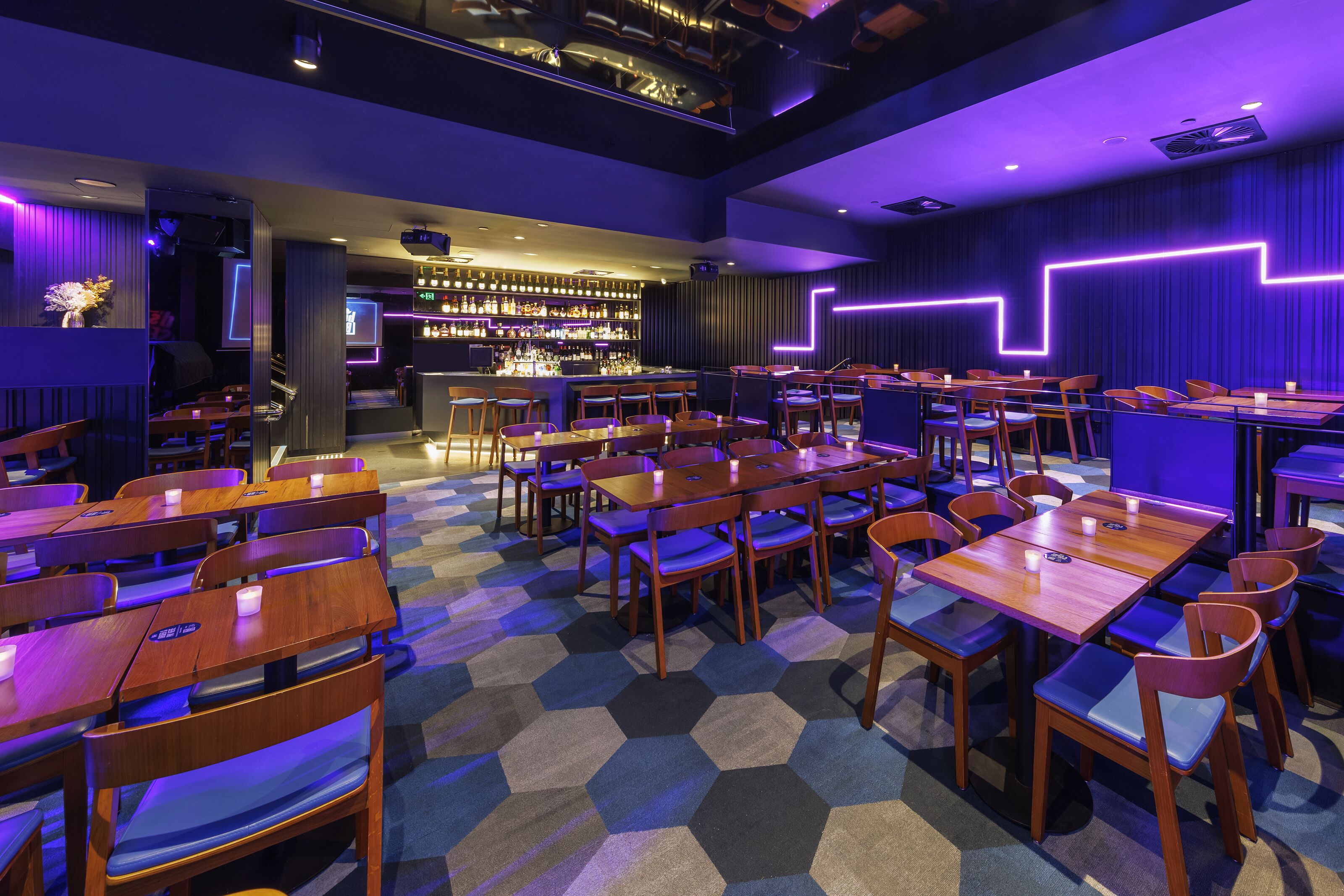 Een restaurant met een hexagonaal patroon in de vloerbedekking, houten tafels en stoelen, en een bar met flessen op de planken.