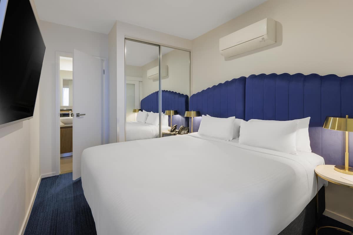 A hotel room with a large bed, a bedside table with a lamp, a mirror, and an air conditioning unit on the wall.