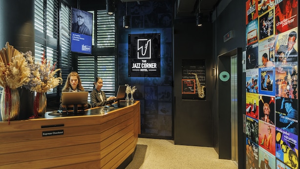 Um balcão de recepção com uma placa indicando "The Jazz Corner Hotel", uma tela de televisão e uma parede decorada com cartazes de filmes.