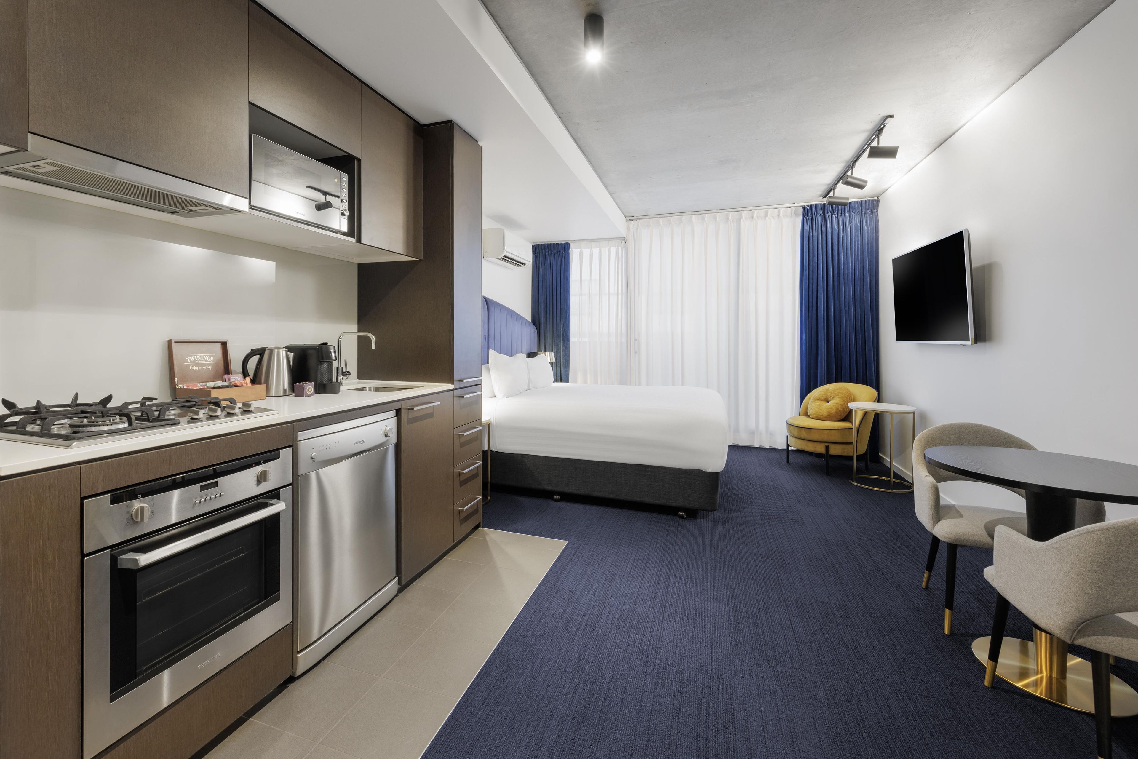 A modern hotel room with a kitchenette, a bed, a flat-screen TV, and a seating area.