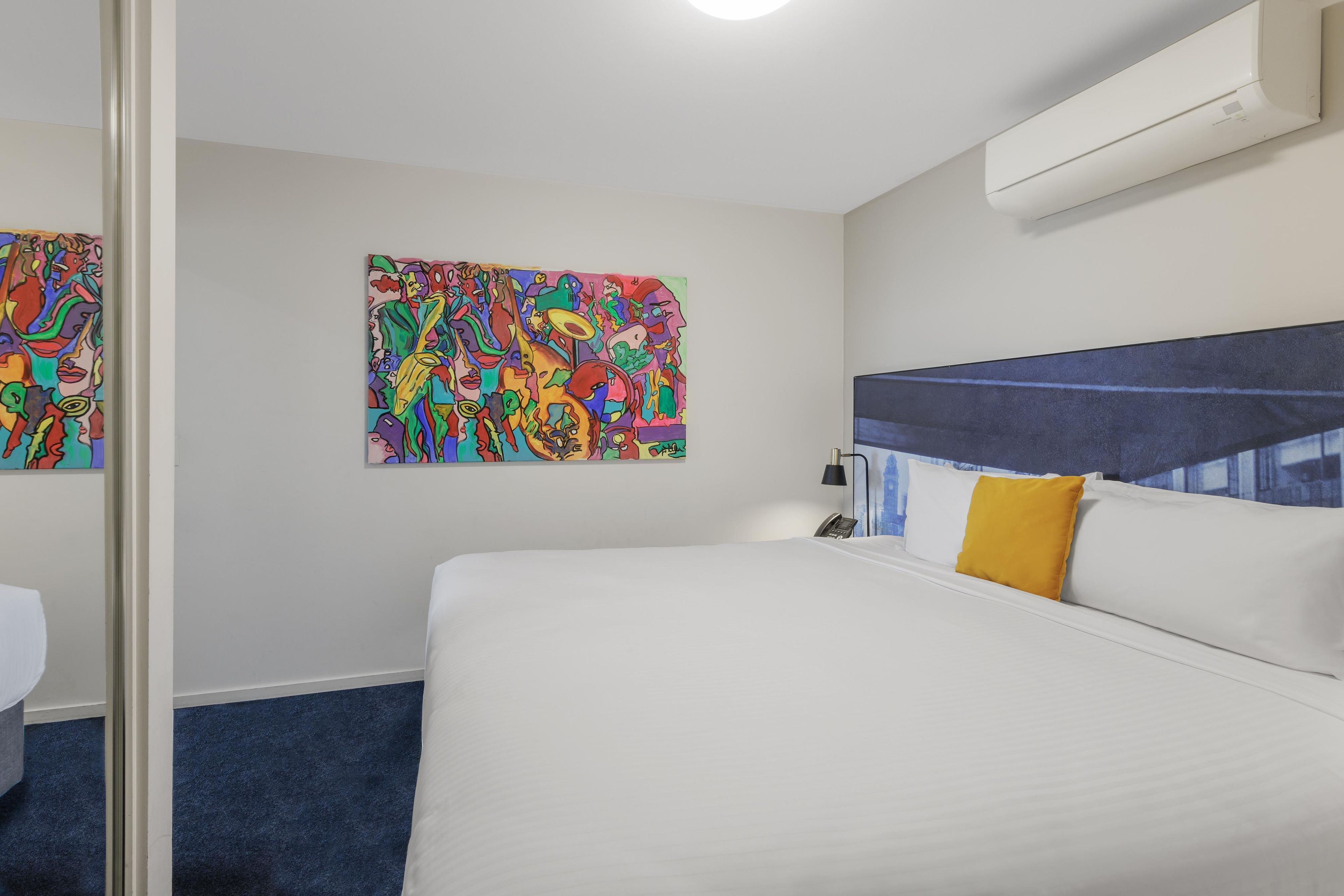 A hotel room with a bed, a wall-mounted air conditioner, a telephone, and two abstract paintings.