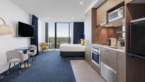 A modern hotel room with a bed, a TV, a desk, a chair, a kitchen area, and a large window offering a city view. - Avani Melbourne Jazz Corner Residences (Melbourne)