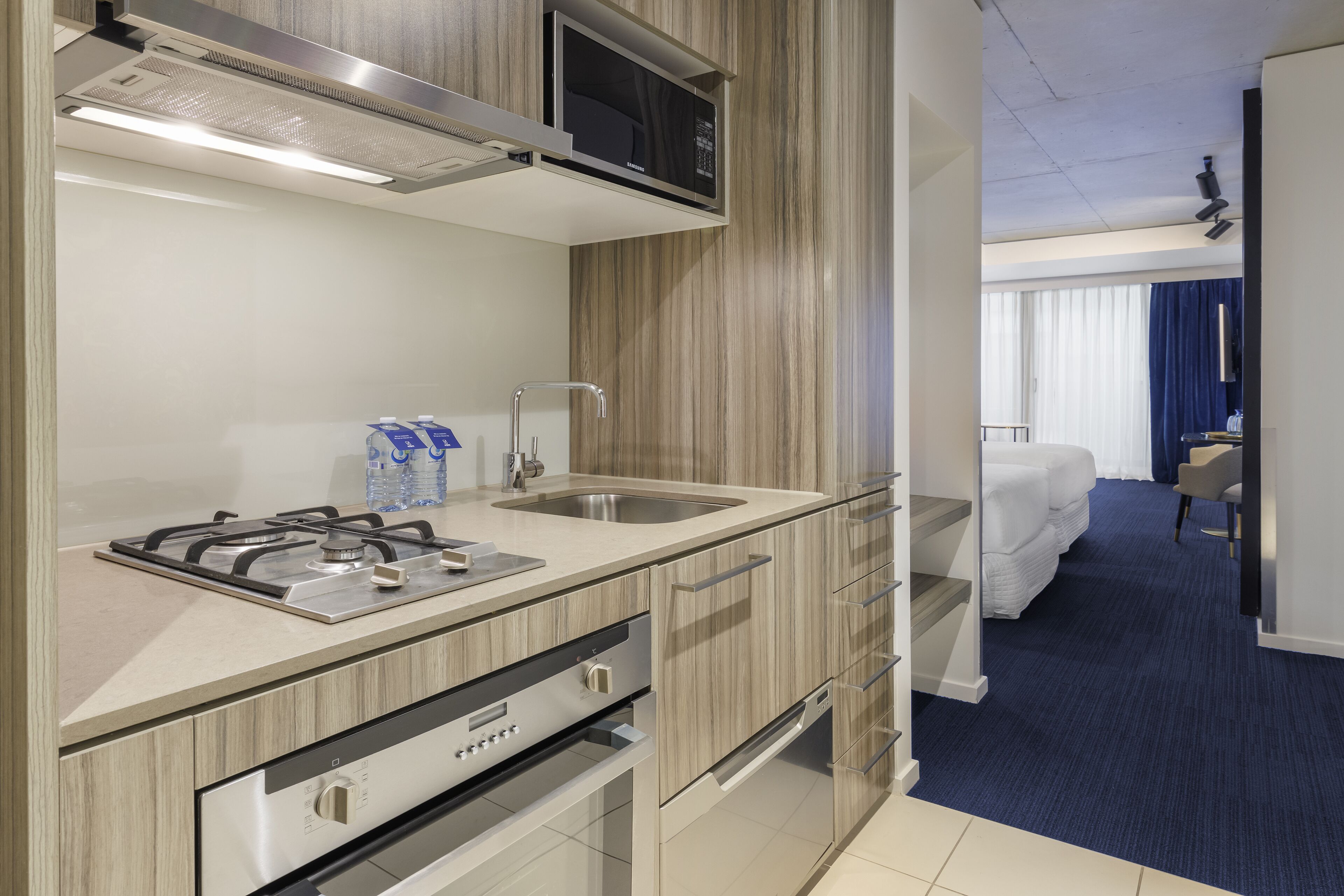 A modern kitchenette with a stove, sink, and microwave. There is a water dispenser and a small refrigerator. The room has wooden cabinetry and a blue carpet.