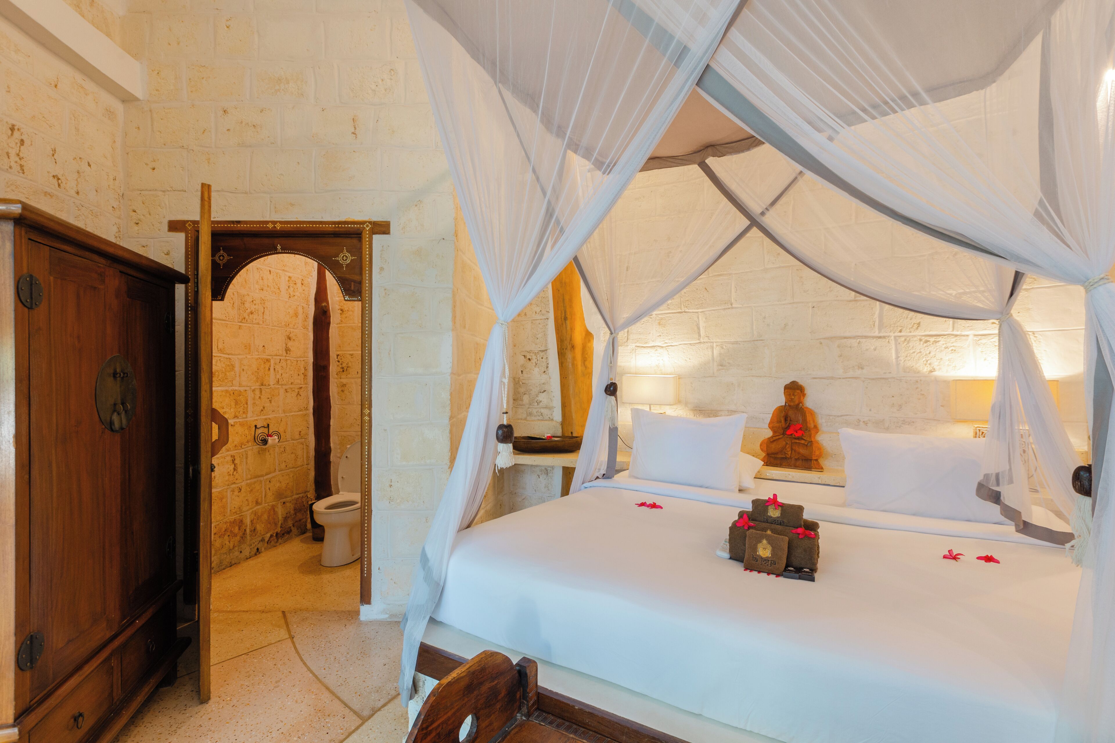 A bedroom with a four-poster bed, a wooden wardrobe, a stone wall, and a Buddha statue.